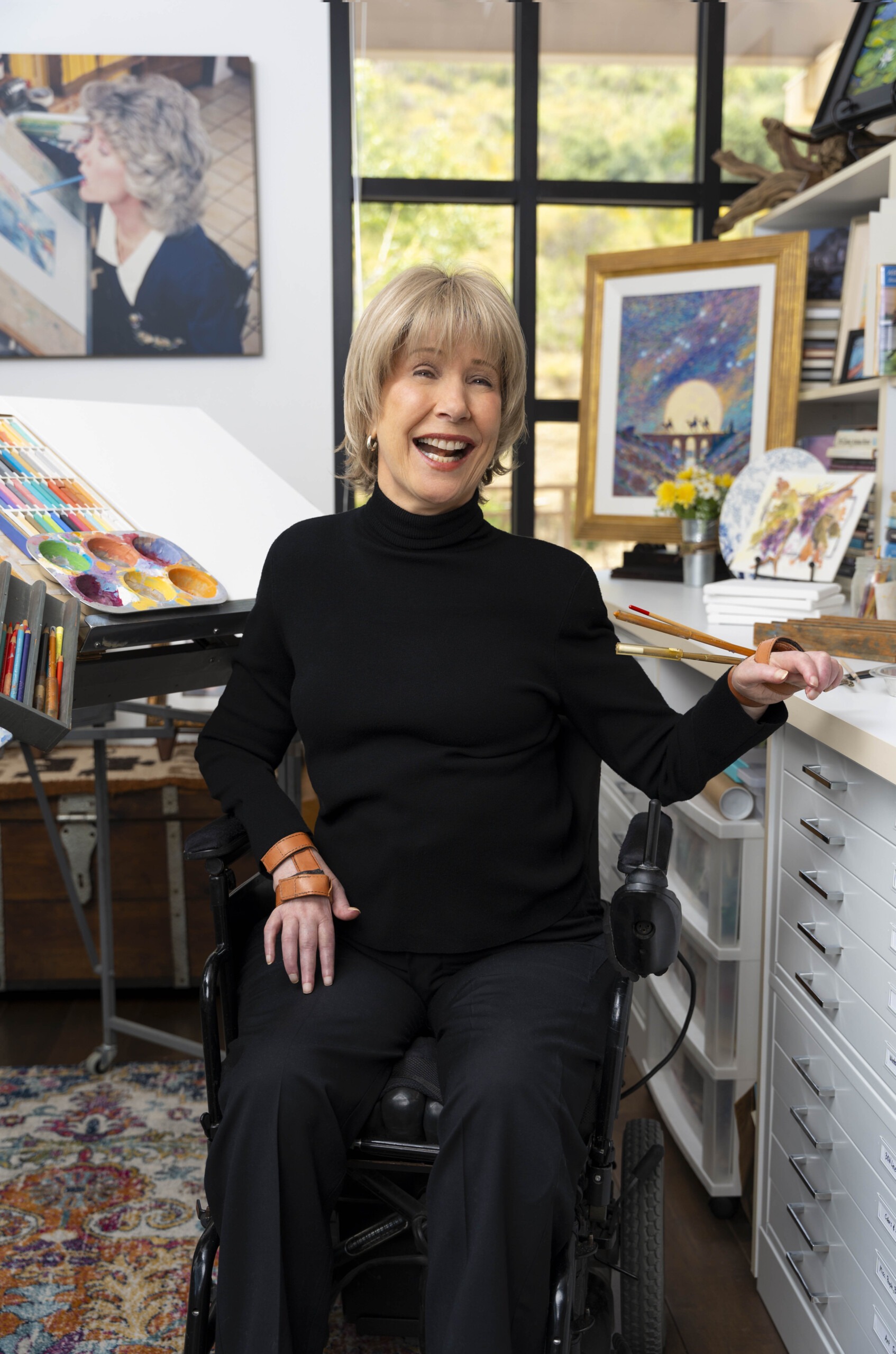 Joni smiling and posing in her art room, with her artwork and art supplies displayed in the background.