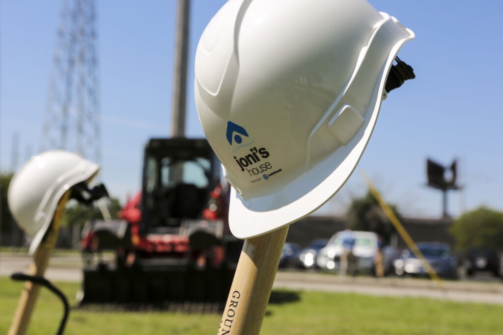 A hard hat helmet branded with the Joni's House logo resting on top of a shovel handle with a large excavator parked in the background.