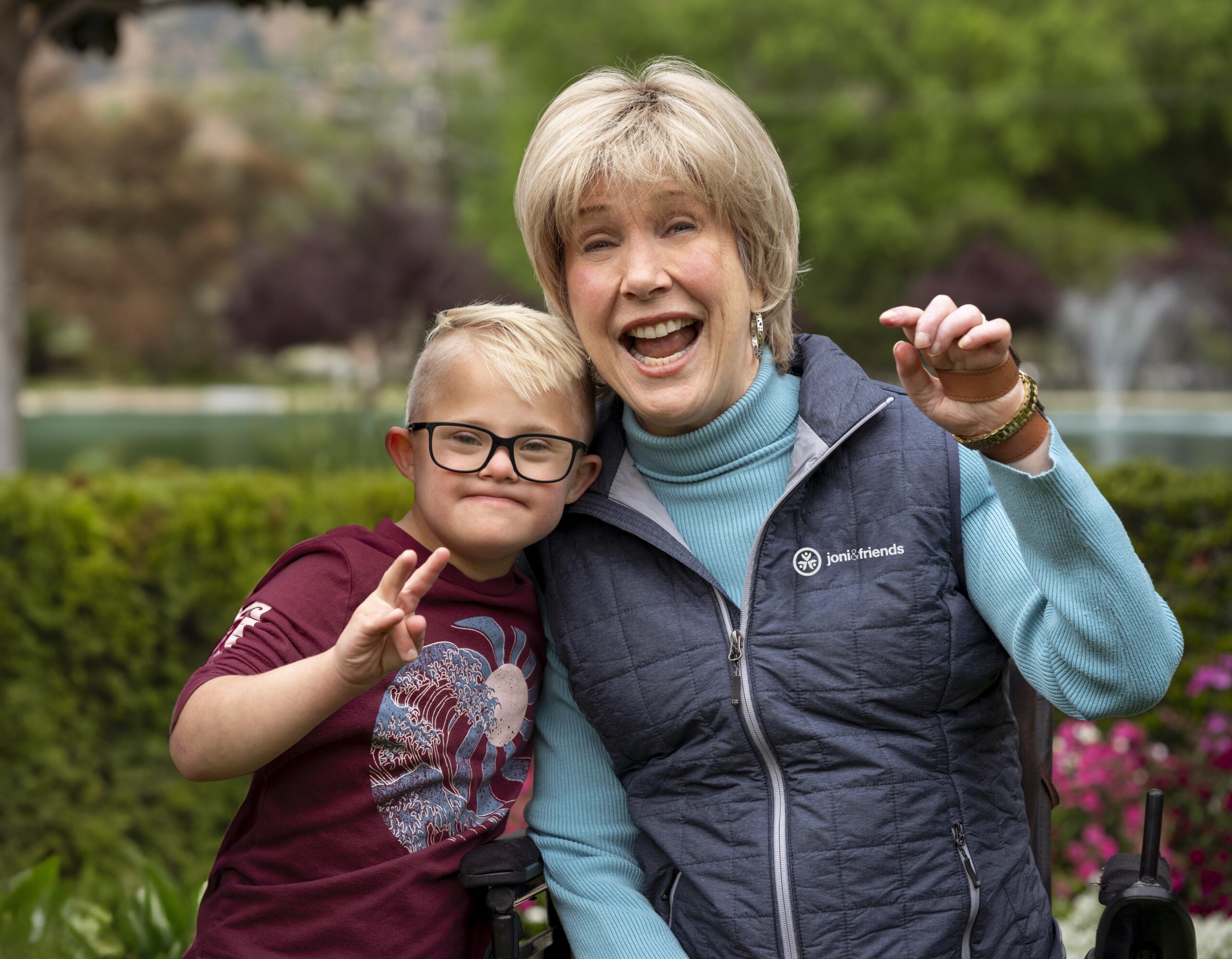 Joni Eareckson Tada seated in her wheelchair smiling at the camera. A young boy with Down syndrome stands next to her displaying the peace sign with his right hand.