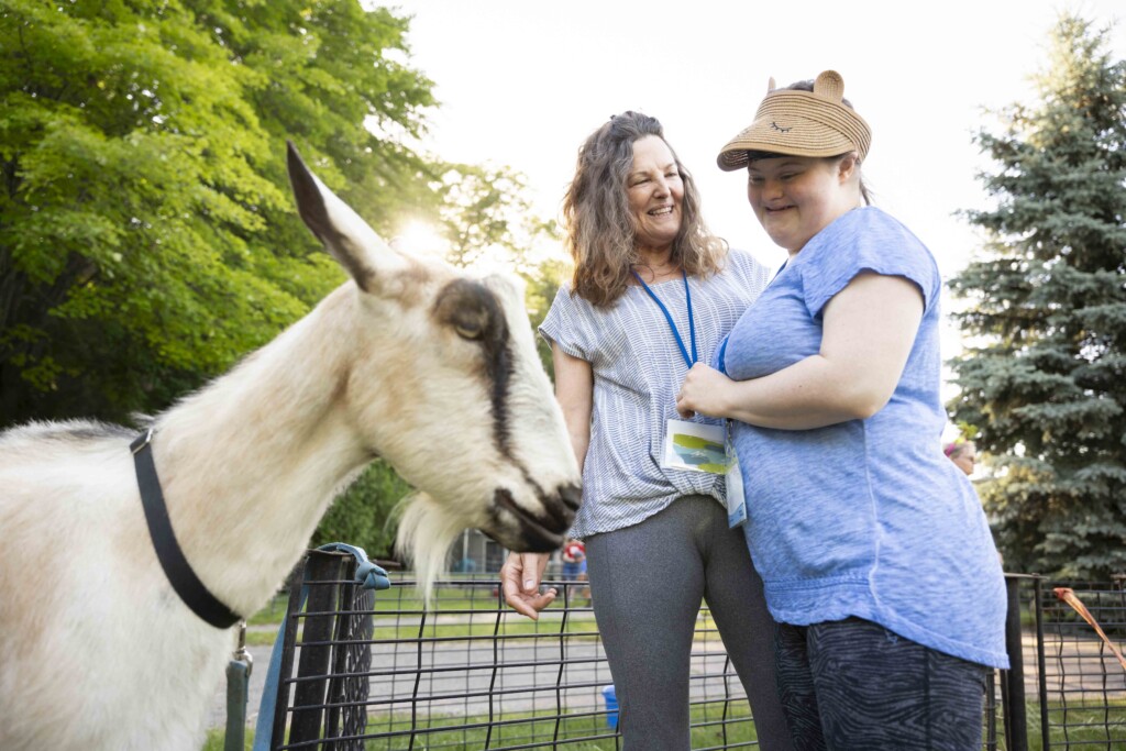 Randi and Pat standing next to each other, both smiling. Randi is looking at a goat.