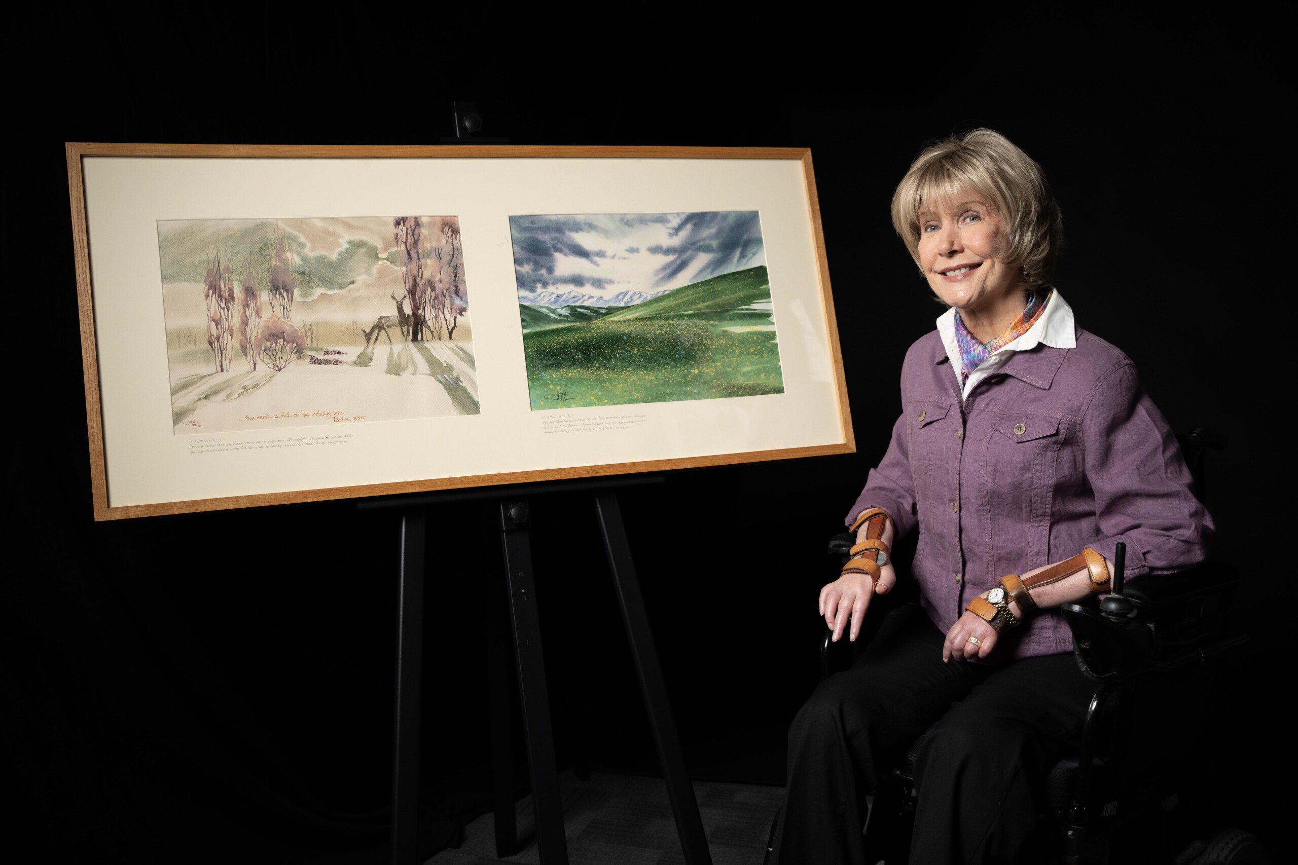 Joni is smiling and sitting in her wheelchair next to her paintings titled "Silent Witness" and "Higher Ground."