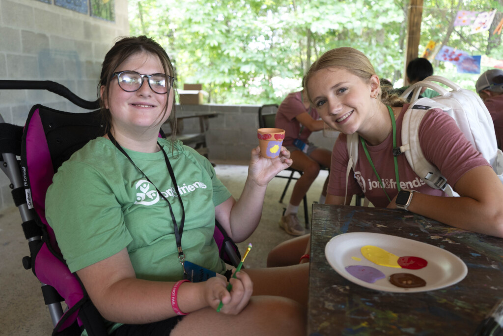 Huntleigh and a volunteer at Joni and Friends both looking at and smiling at the camera while Huntleigh paints a small plant pot.