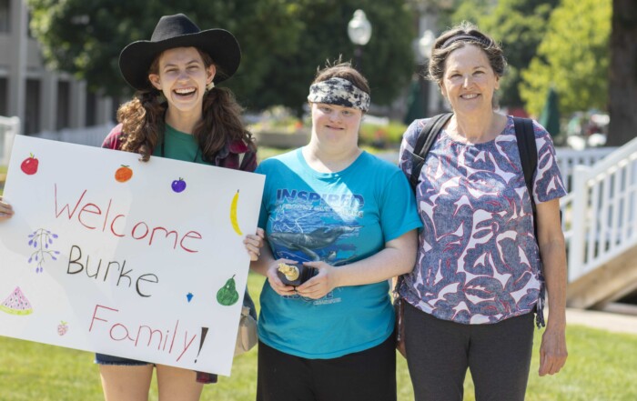 Kate holding a colorful "Welcome Burke Family" sign. Randi and Pat standing beside her, all three smiling at the camera.