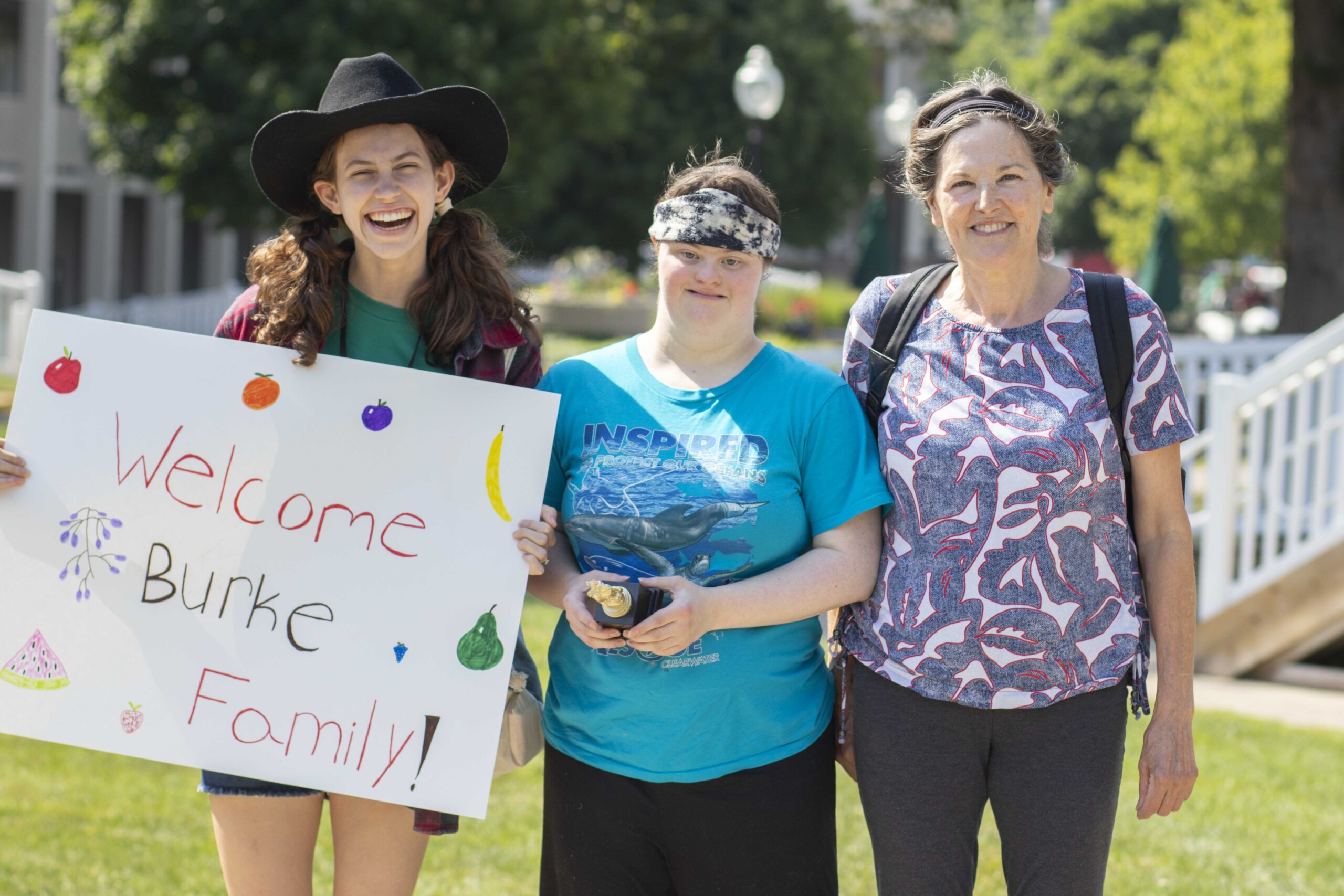 Kate holding a colorful "Welcome Burke Family" sign. Randi and Pat standing beside her, all three smiling at the camera.