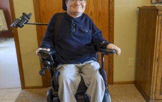 Joel Vander Molen seated in his wheelchair, smiling directly at the camera in a full-body shot.