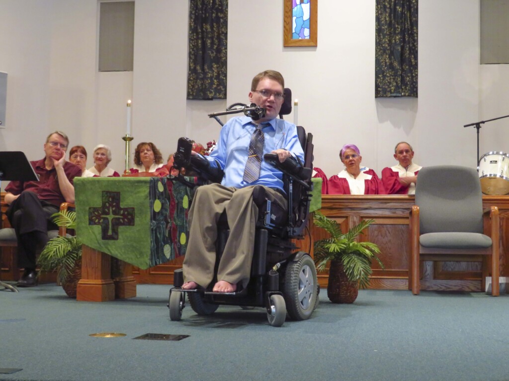 Joel Vander Molen seated in his wheelchair, speaking at a church, shown in a full-body shot.