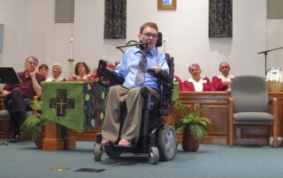 Joel Vander Molen seated in his wheelchair, speaking at a church, shown in a full-body shot.