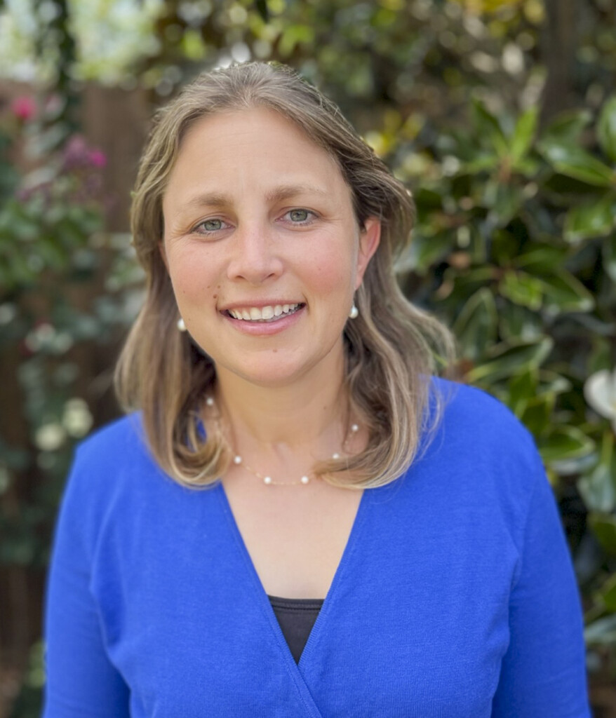 A half-body shot of Rachel smiling at the camera with plants in the background.