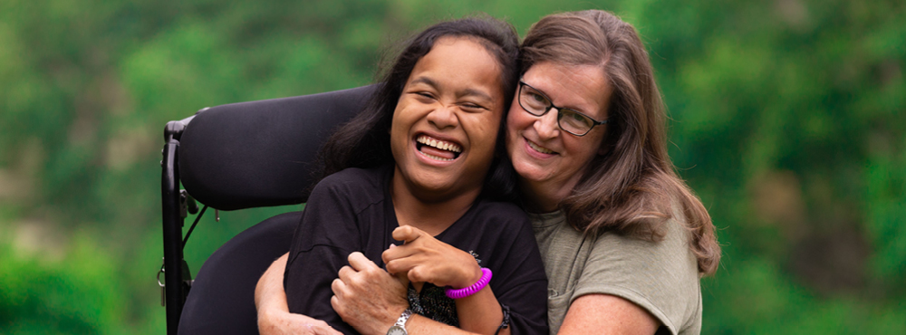 A woman and a girl in a wheelchair are smiling happily at the camera while hugging each other.