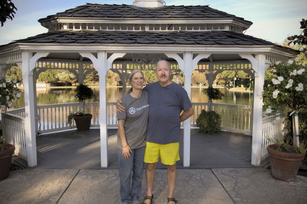 Kathy and Mike are standing next to each other while smiling and hugging each other with a lake visible in the background.