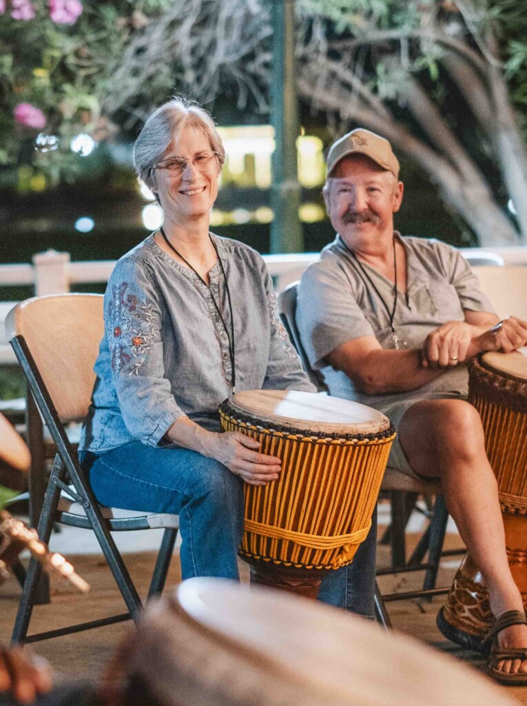 Kathy and Mike are smiling and sitting next to each other, each holding a djembe drum, while looking at another drummer.