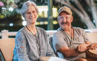 Kathy and Mike are smiling and sitting next to each other, each holding a djembe drum, while looking at another drummer.