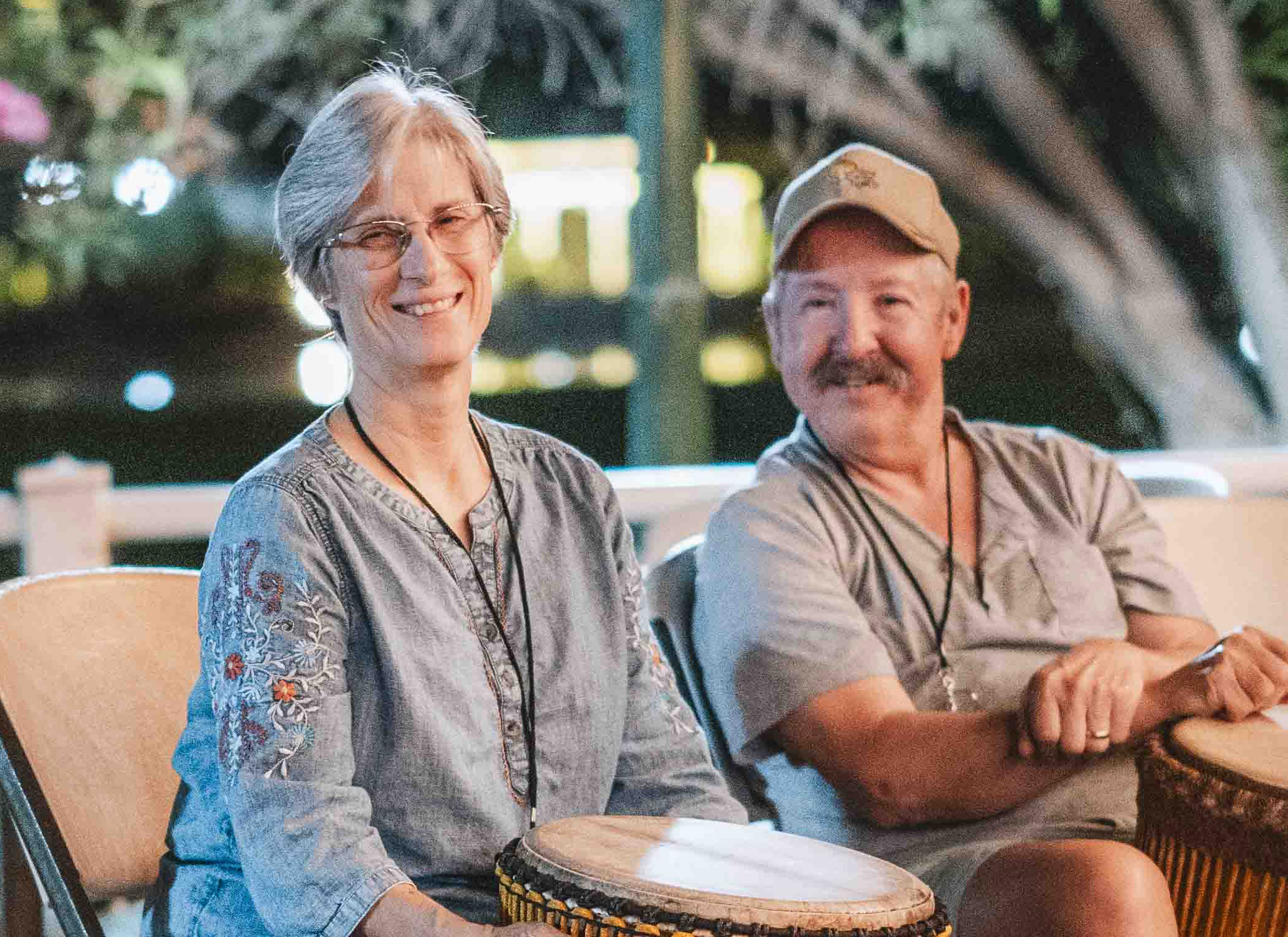 Kathy and Mike are smiling and sitting next to each other, each holding a djembe drum, while looking at another drummer.