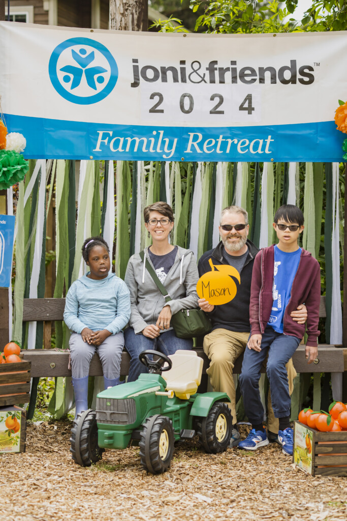 Becky and Nathan with their two youngest children, Lydia and Caleb, sitting and smiling at the camera, with a "Joni and Friends 2024 Family Retreat" banner in the background.