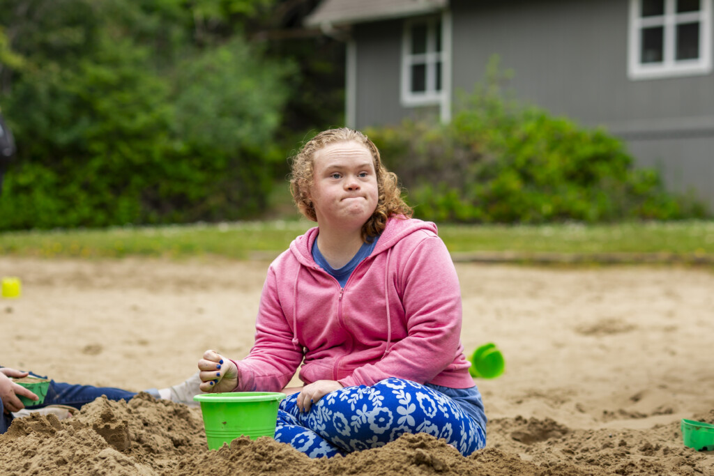 Colleen is sitting on the sand with a green bucket in front of her.