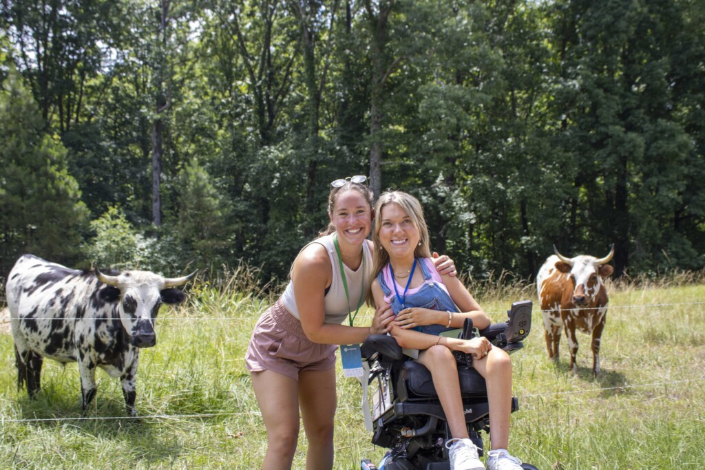Lindsay Cochran sits in her wheelchair, with an intern gently holding her shoulders. They are outdoors with a natural backdrop, where two Pineywoods cattle can be seen in the background—one with black and white spots and the other with brown and white spots.