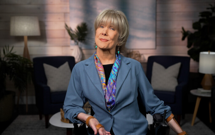 Joni Eareckson Tada smiling and posing for the camera, wearing a blue blazer, a colorful scarf, and blue earrings. The background is a cozy indoor living room setting.