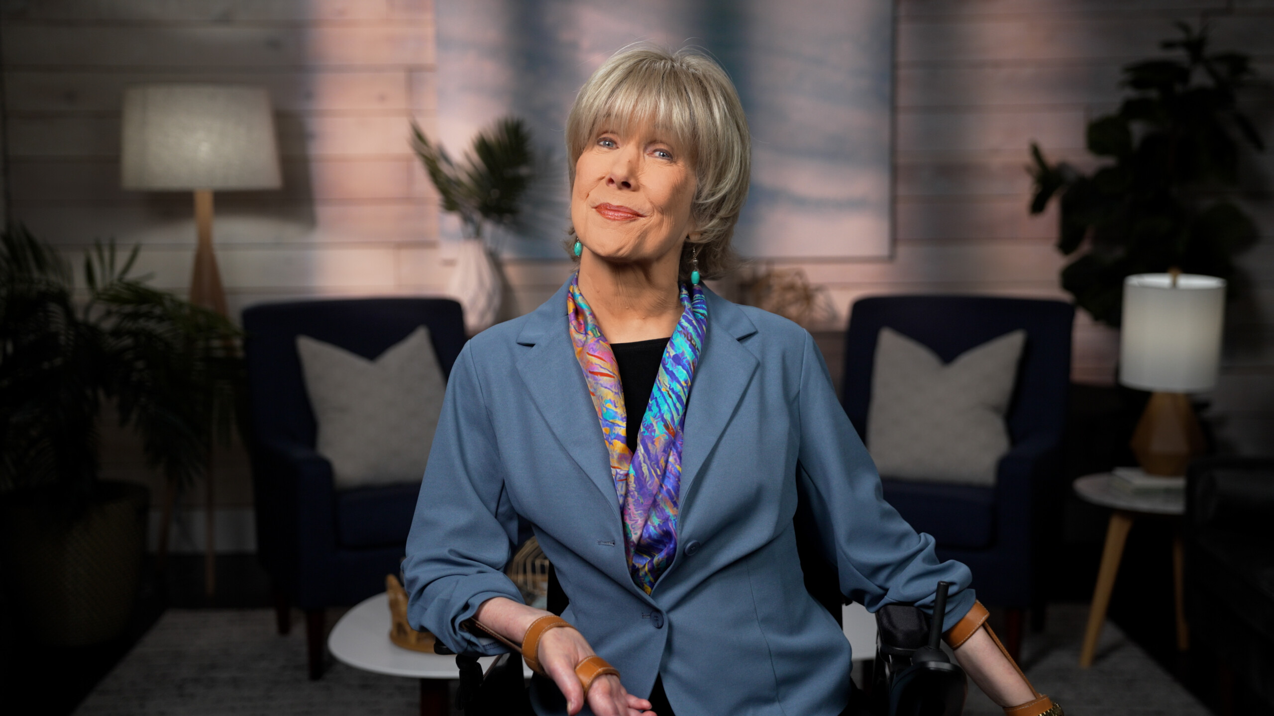 Joni Eareckson Tada smiling and posing for the camera, wearing a blue blazer, a colorful scarf, and blue earrings. The background is a cozy indoor living room setting.