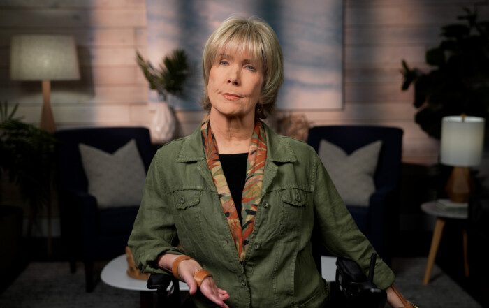 Joni Eareckson Tada, wearing a green denim jacket and a scarf, is looking at the camera. The background features a living room setting.