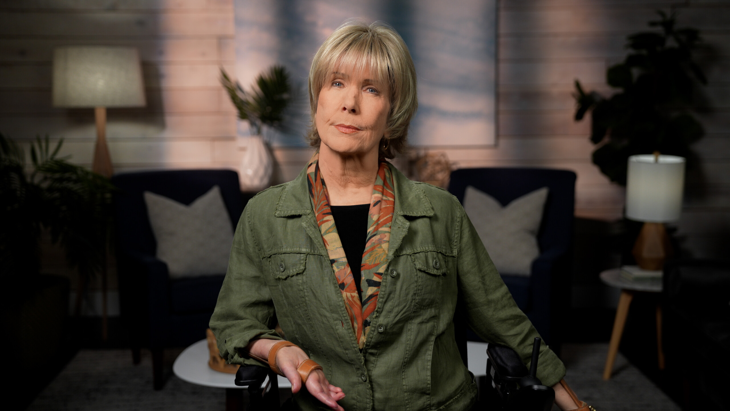 Joni Eareckson Tada, wearing a green denim jacket and a scarf, is looking at the camera. The background features a living room setting.