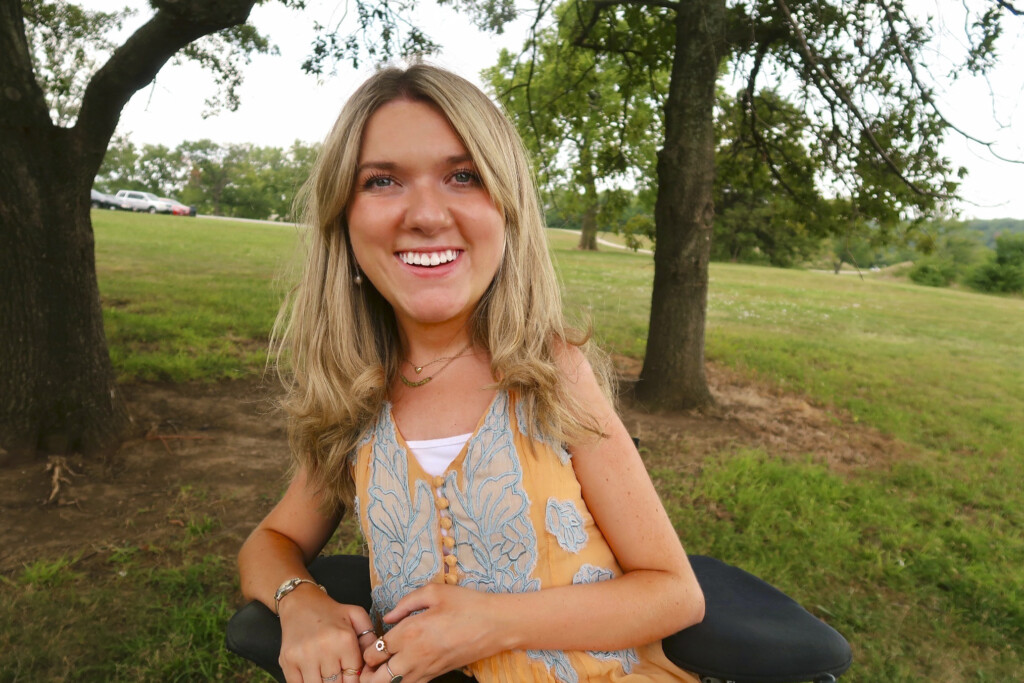 Lindsay Cochran smiles warmly at the camera against a lush background of green grass and trees, radiating happiness and positivity.