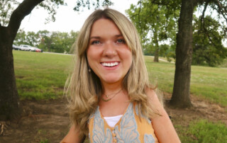 Lindsay Cochran smiles warmly at the camera against a lush background of green grass and trees, radiating happiness and positivity.