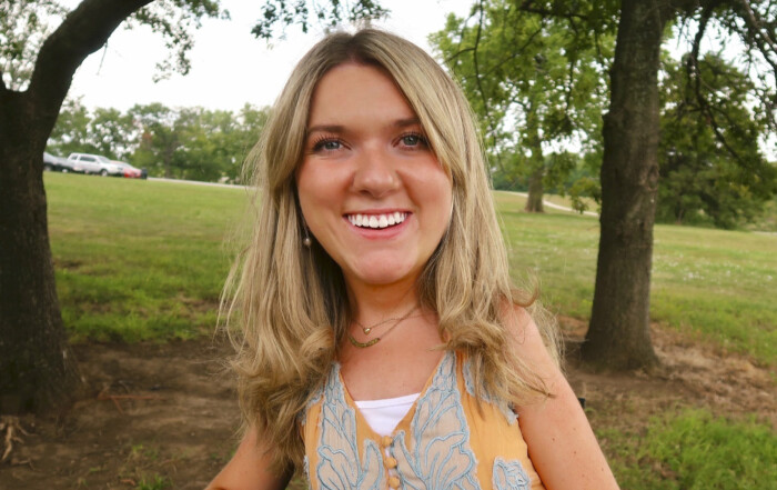 Lindsay Cochran smiles warmly at the camera against a lush background of green grass and trees, radiating happiness and positivity.