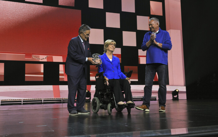 Joni Eareckson Tada on stage with Ken standing beside her, holding her GLS award. Another man next to them is clapping in celebration.