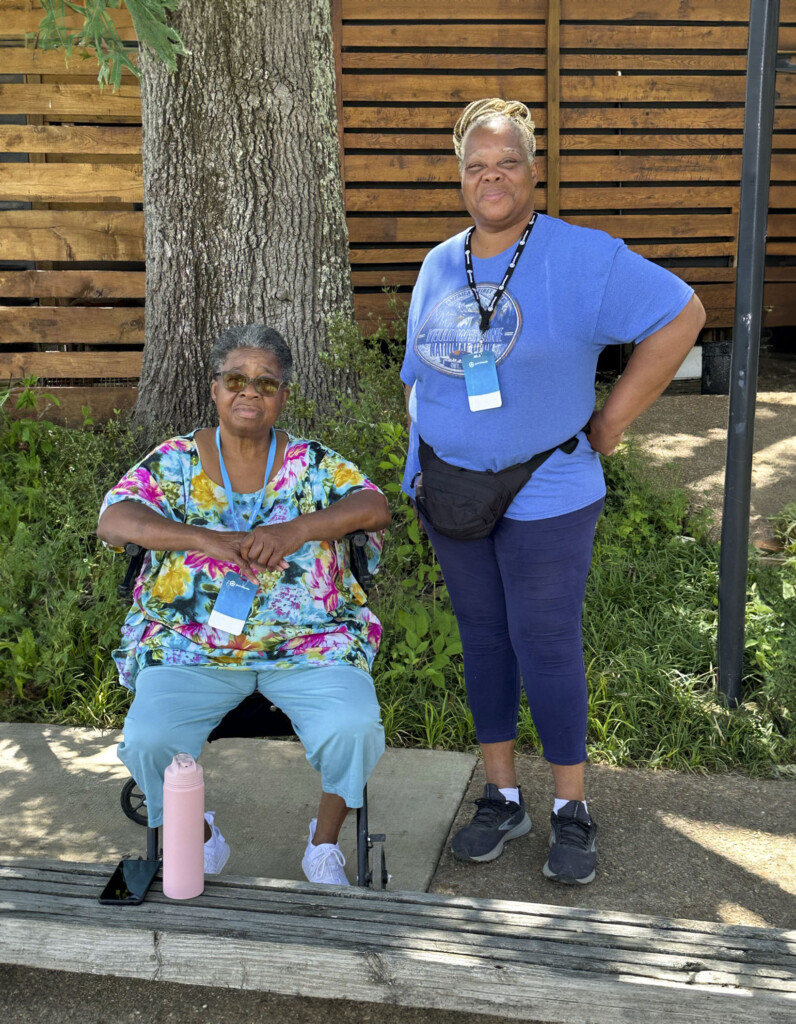 Shirley and Maxine are next to each other and posing for the camera outdoors, under the shade of a tree.