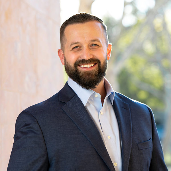Jason Holden smiles confidently at the camera, dressed in a formal suit against a softly blurred tree background.