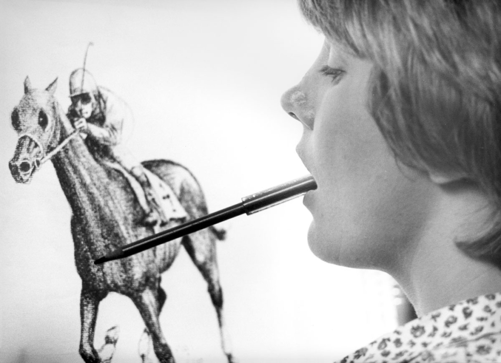 A black-and-white close-up side view of Joni Eareckson Tada, who is using a pencil held in her mouth to draw. She is focused on sketching a horse and a rider on the paper.