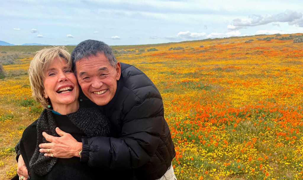 A picture of Ken and Joni embracing each other with joy. Both are smiling widely, and they are surrounded by a vibrant, colorful field of flowers in the background.