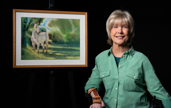 Joni, dressed in a vibrant green button-up long sleeve shirt, smiles at the camera beside her captivating painting titled "Horse in Wild Mustard." The artwork, rich in color and detail, features a majestic horse set against a backdrop of wild mustard flowers, highlighting her artistic talent and passion.