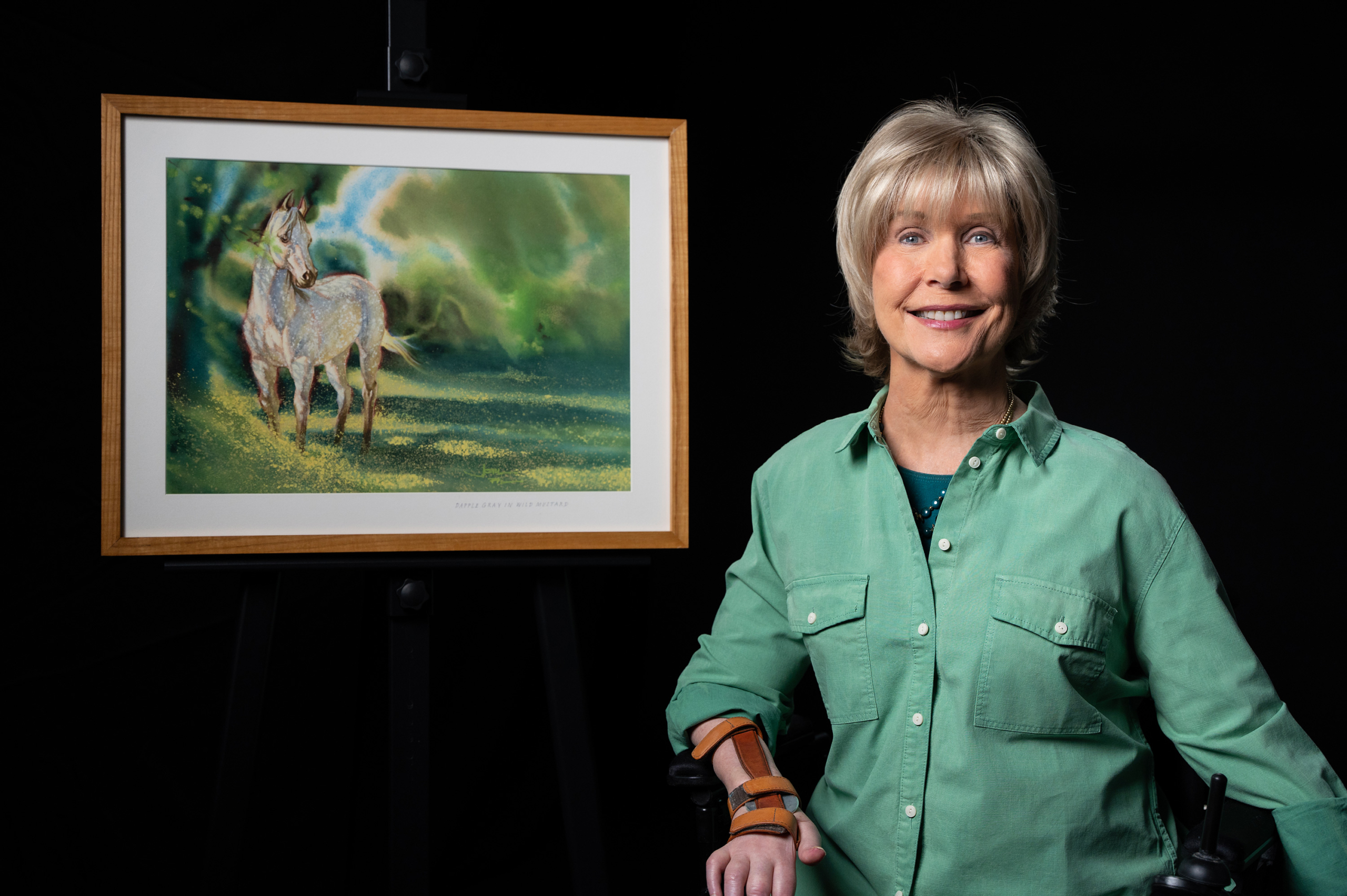 Joni, dressed in a vibrant green button-up long sleeve shirt, smiles at the camera beside her captivating painting titled "Horse in Wild Mustard." The artwork, rich in color and detail, features a majestic horse set against a backdrop of wild mustard flowers, highlighting her artistic talent and passion.