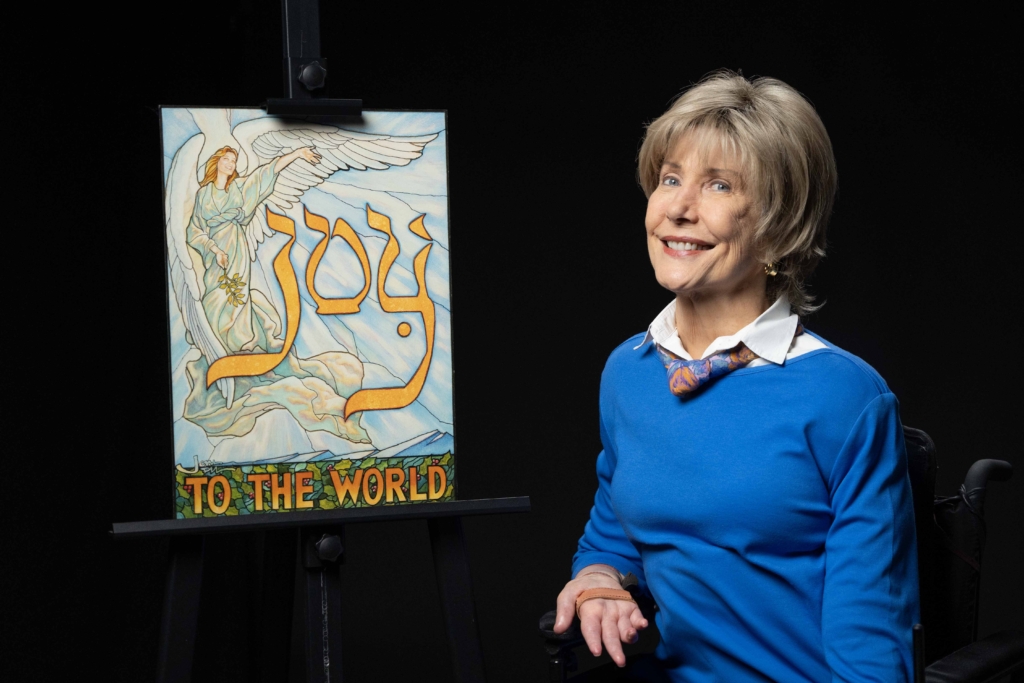 Joni wearing a bright blue long sleeve shirt with a colorful scarf, smiling at the camera next to her "Joy to the World" artwork. 