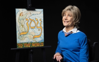 Joni wearing a bright blue long sleeve with a colorful scarf smiling at the camera next to her "Joy to the World" artwork.