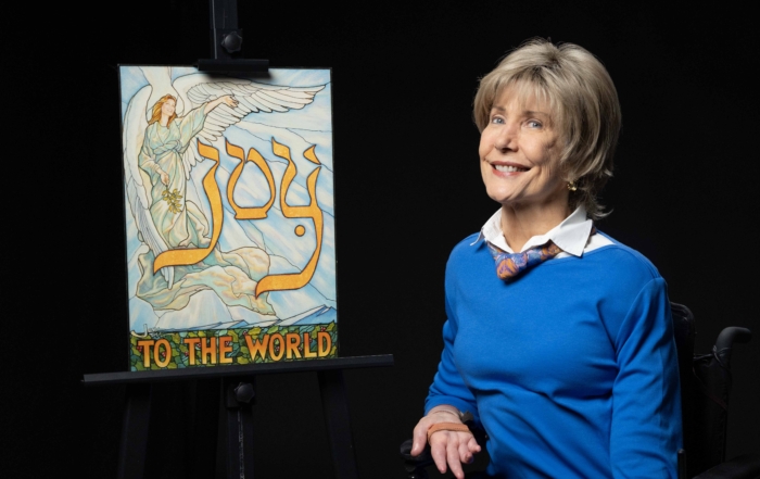 Joni wearing a bright blue long sleeve with a colorful scarf smiling at the camera next to her "Joy to the World" artwork.