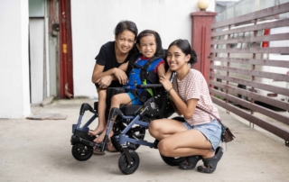 Robby's mom is on the left, Robby is seated in his new Cub wheelchair in the middle, and Patricia, Robby’s sister is on the right, all are posing and smiling for the camera.