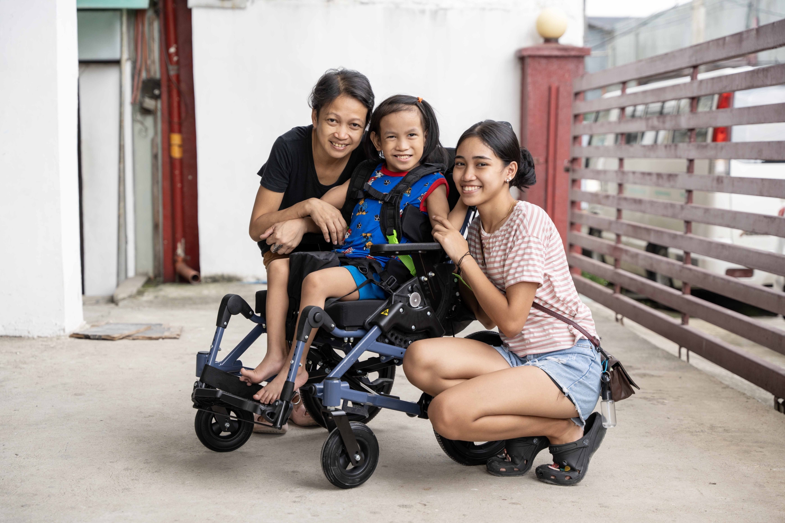 Robby's mom is on the left, Robby is seated in his new Cub wheelchair in the middle, and Patricia, Robby’s sister is on the right, all are posing and smiling for the camera.