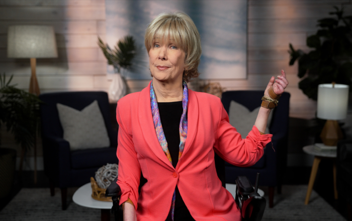 Joni Eareckson Tada, wearing a vibrant light red blazer over a black top and a colorful scarf, is smiling as she raises her left arm for the camera. The background features a softly blurred living room setup, adding a cozy atmosphere to the image.