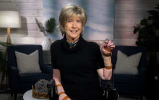 Joni wearing a black blouse and a colorful scarf, smiling and posing for the camera in a cozy living room setup.