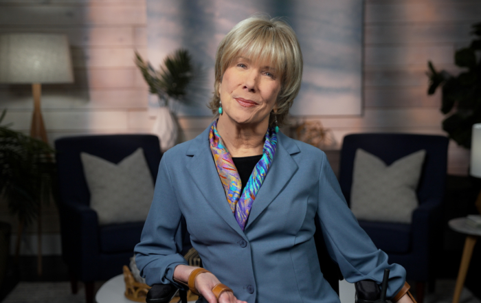 Joni smiling while wearing a blue blazer, a colorful scarf, and turquoise earrings, with a living room background.