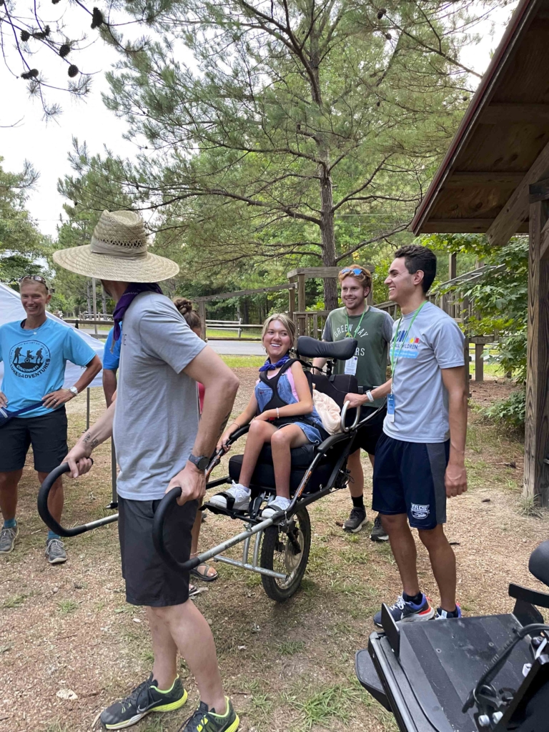 Lindsay Cochran being carried in a specialized wheelchair designed for hiking by volunteers from Luke5Aventures, with the group navigating a trail together.
