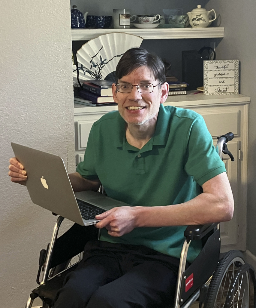 Kevin Avery, a cheerful man in a green polo shirt, is seated in his wheelchair, holding a MacBook laptop. He is smiling brightly at the camera, radiating warmth and positivity.