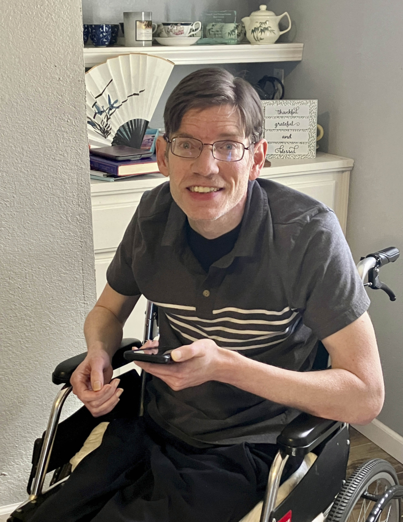 Kevin Avery, a cheerful man in a grey polo shirt with stripes in the middle, is seated in his wheelchair, holding a phone with his left hand. He is smiling brightly at the camera, radiating warmth and positivity.