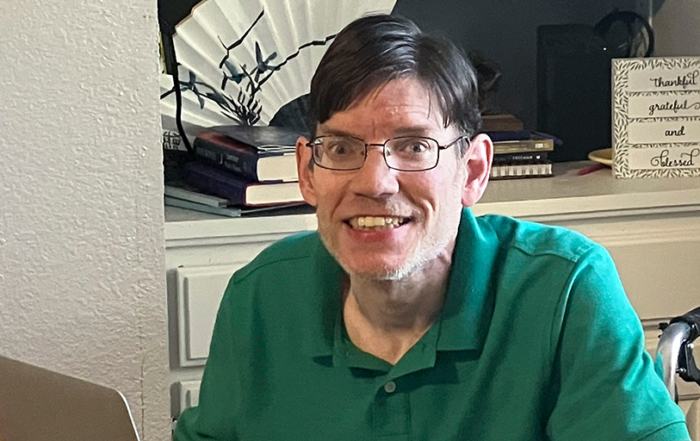 Kevin Avery, a cheerful man in a green polo shirt, is seated in his wheelchair, holding a MacBook laptop. He is smiling brightly at the camera, radiating warmth and positivity.