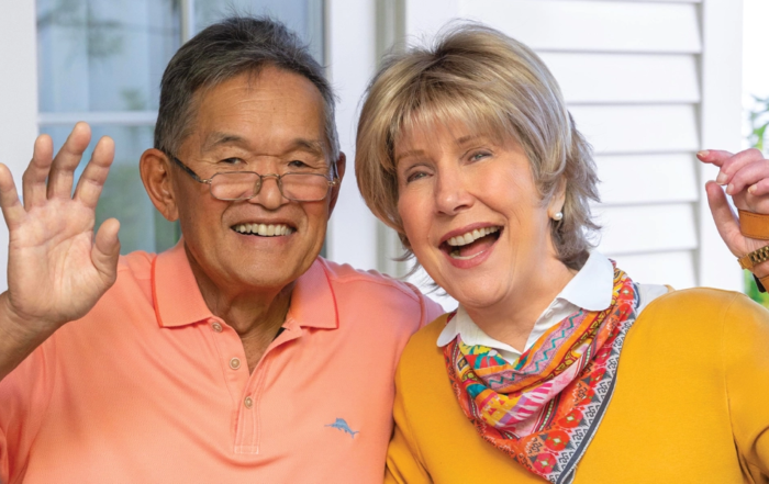 Ken, on the right, is wearing a bright orange polo shirt, while Joni is dressed in a bright yellow top with half-length sleeves and a colorful festive scarf. Both Ken and Joni are smiling and waving at the camera.