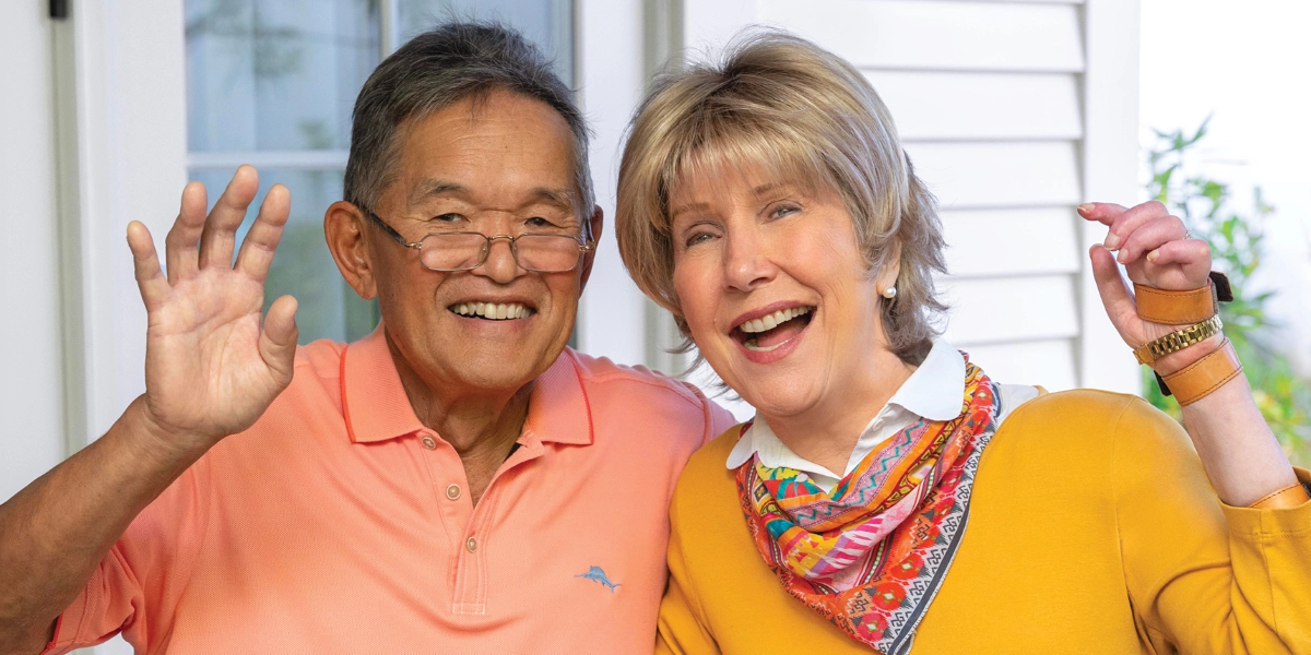 Ken, on the right, is wearing a bright orange polo shirt, while Joni is dressed in a bright yellow top with half-length sleeves and a colorful festive scarf. Both Ken and Joni are smiling and waving at the camera.