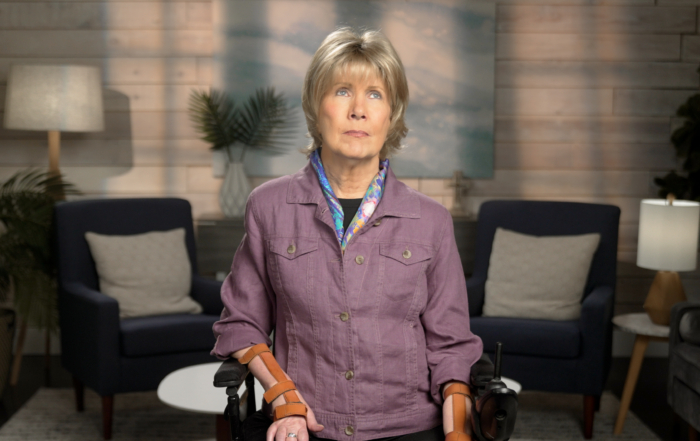 Joni wearing a purple cardigan jacket and a colorful scarf against a living room background.