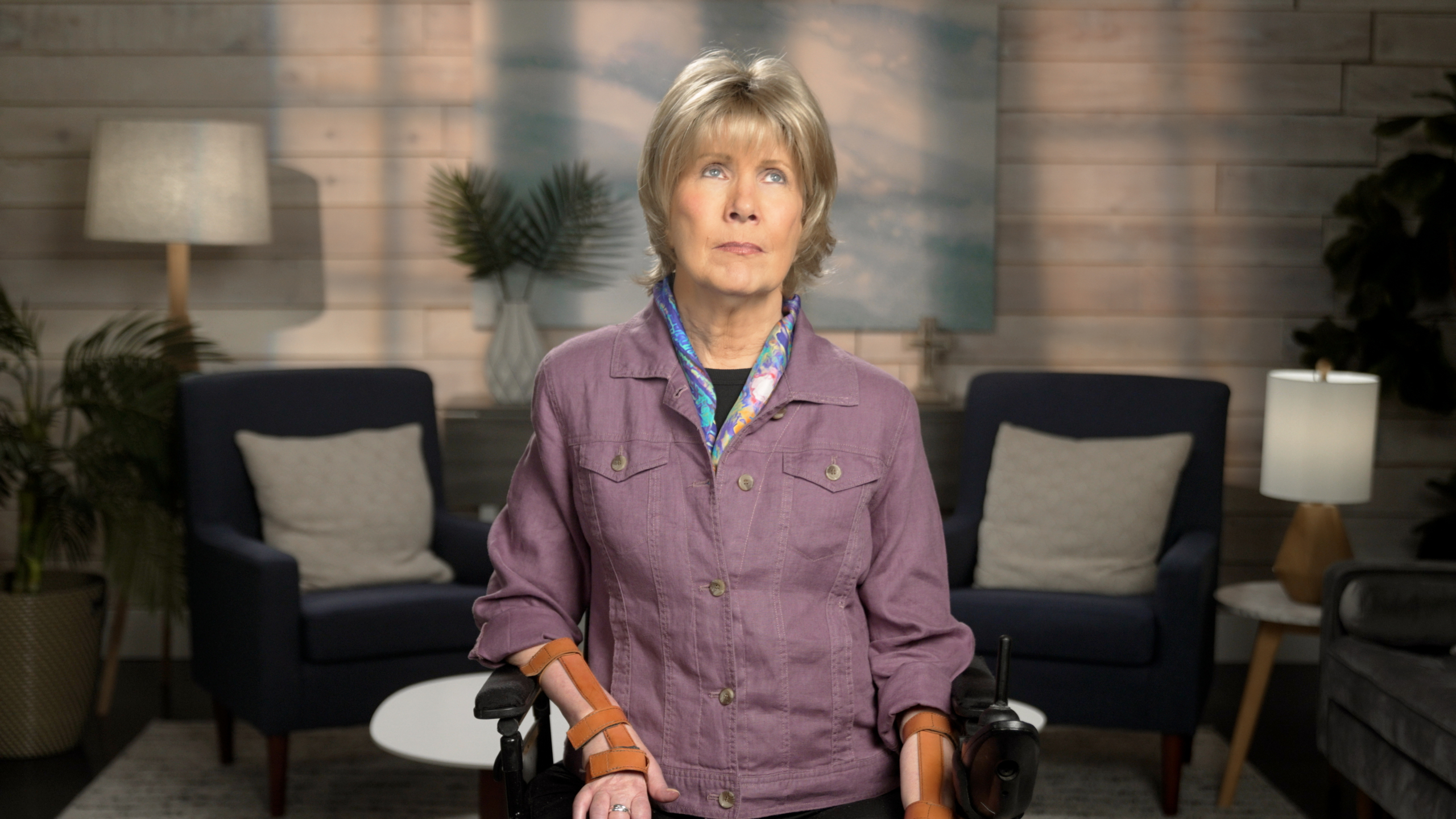 Joni wearing a purple cardigan jacket and a colorful scarf against a living room background.